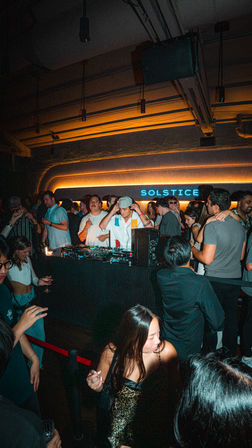 Energetic urban nightclub scene with a DJ at the booth, a packed dancing crowd under warm neon backlighting, and a woman in a leopard-print dress in the foreground.