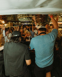 Two DJs playing to a lively riverfront boat party at night, crowd dancing under a glowing city skyline and bridge