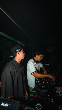 Two DJs in a dark nightclub DJ booth — one with a backwards cap and headphones, the other adjusting a multi-deck mixer with colorful LED lights, late-night urban party vibe.