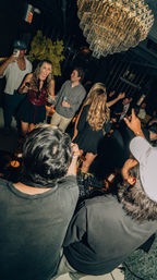 Energetic nightclub lounge scene with DJs at a booth and a dancing crowd holding drinks and taking photos beneath a large crystal chandelier.
