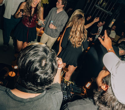 Energetic nightclub lounge scene with DJs at a booth and a dancing crowd holding drinks and taking photos beneath a large crystal chandelier.
