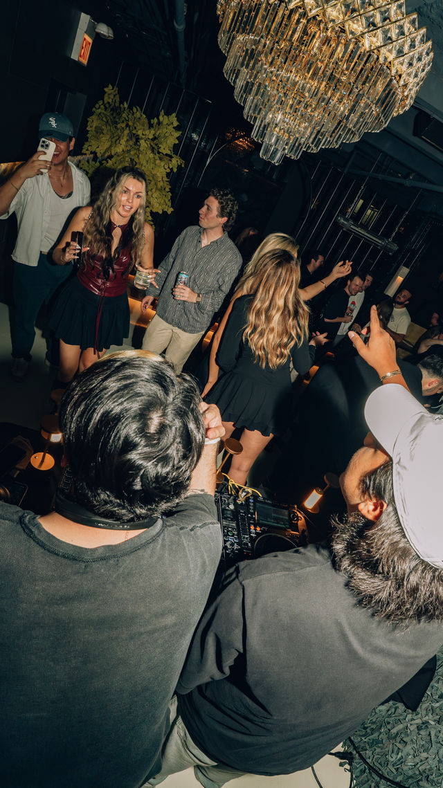Energetic nightclub lounge scene with DJs at a booth and a dancing crowd holding drinks and taking photos beneath a large crystal chandelier.