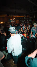 DJ in a white jacket and cap spinning at a crowded city nightclub DJ booth, lively dance floor, bar shelves with bottles, neon sign and patrons with drinks and phones.