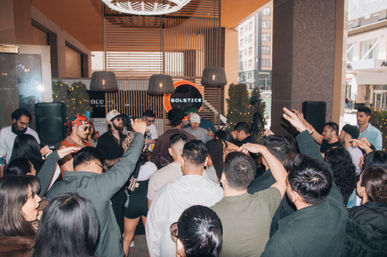 Lively urban venue party with a DJ booth and neon sign, crowd of people dancing and cheering with hands in the air