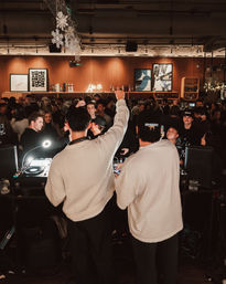 Two DJs at a packed indoor bar DJ booth energizing a lively nightlife crowd under hanging snowflake decorations and a neon sign above a wood-paneled back bar.