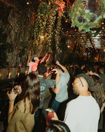 Energetic crowd dancing at a jungle-themed urban patio bar with a DJ booth, hanging vines, disco balls and warm string lights for lively nightlife