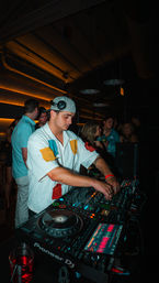 Focused DJ in a backwards cap mixing on Pioneer DJ decks in a dim, warm-lit nightclub with a lively crowd and drinks in the background