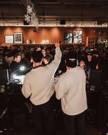 Two DJs at a packed indoor bar DJ booth energizing a lively nightlife crowd under hanging snowflake decorations and a neon sign above a wood-paneled back bar.