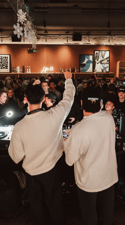 Two DJs at a packed indoor bar DJ booth energizing a lively nightlife crowd under hanging snowflake decorations and a neon sign above a wood-paneled back bar.