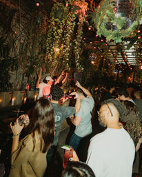 Energetic crowd dancing at a jungle-themed urban patio bar with a DJ booth, hanging vines, disco balls and warm string lights for lively nightlife