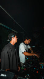 Two DJs in a dark nightclub DJ booth — one with a backwards cap and headphones, the other adjusting a multi-deck mixer with colorful LED lights, late-night urban party vibe.