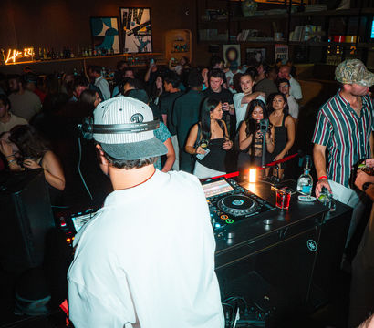 DJ in a white jacket and cap spinning at a crowded city nightclub DJ booth, lively dance floor, bar shelves with bottles, neon sign and patrons with drinks and phones.