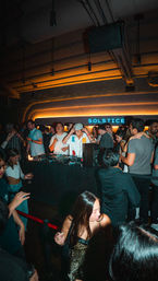Energetic urban nightclub scene with a DJ at the booth, a packed dancing crowd under warm neon backlighting, and a woman in a leopard-print dress in the foreground.
