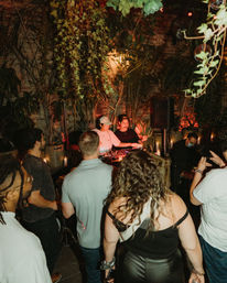 Crowd enjoying a lively evening DJ set at a plant-filled urban garden bar with hanging vines, warm candlelight, and tropical decor.