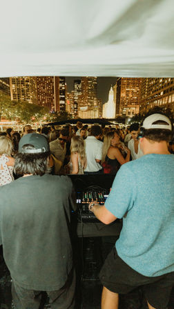 Nighttime boat party with two DJs mixing for a packed crowd, lively people dancing with an illuminated downtown skyline in the background.