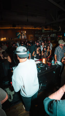 DJ in a white jacket and cap spinning at a crowded city nightclub DJ booth, lively dance floor, bar shelves with bottles, neon sign and patrons with drinks and phones.