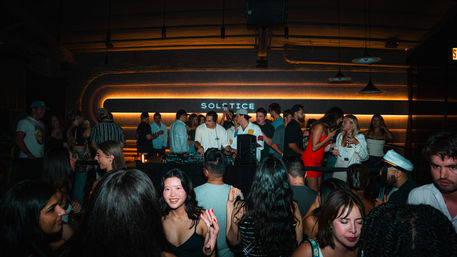 Packed urban nightclub with a DJ at a neon-backlit booth and young adults dancing and socializing under warm amber lights.
