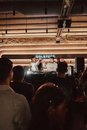 Neon-lit indoor DJ set in a warm modern venue — two DJs fist-bumping behind turntables as a crowd watches under ambient overhead lighting and exposed ceiling details
