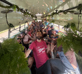 Happy group on a greenery-decorated party bus during a daytime road trip on a rural road, people smiling, holding drinks and posing for a selfie.