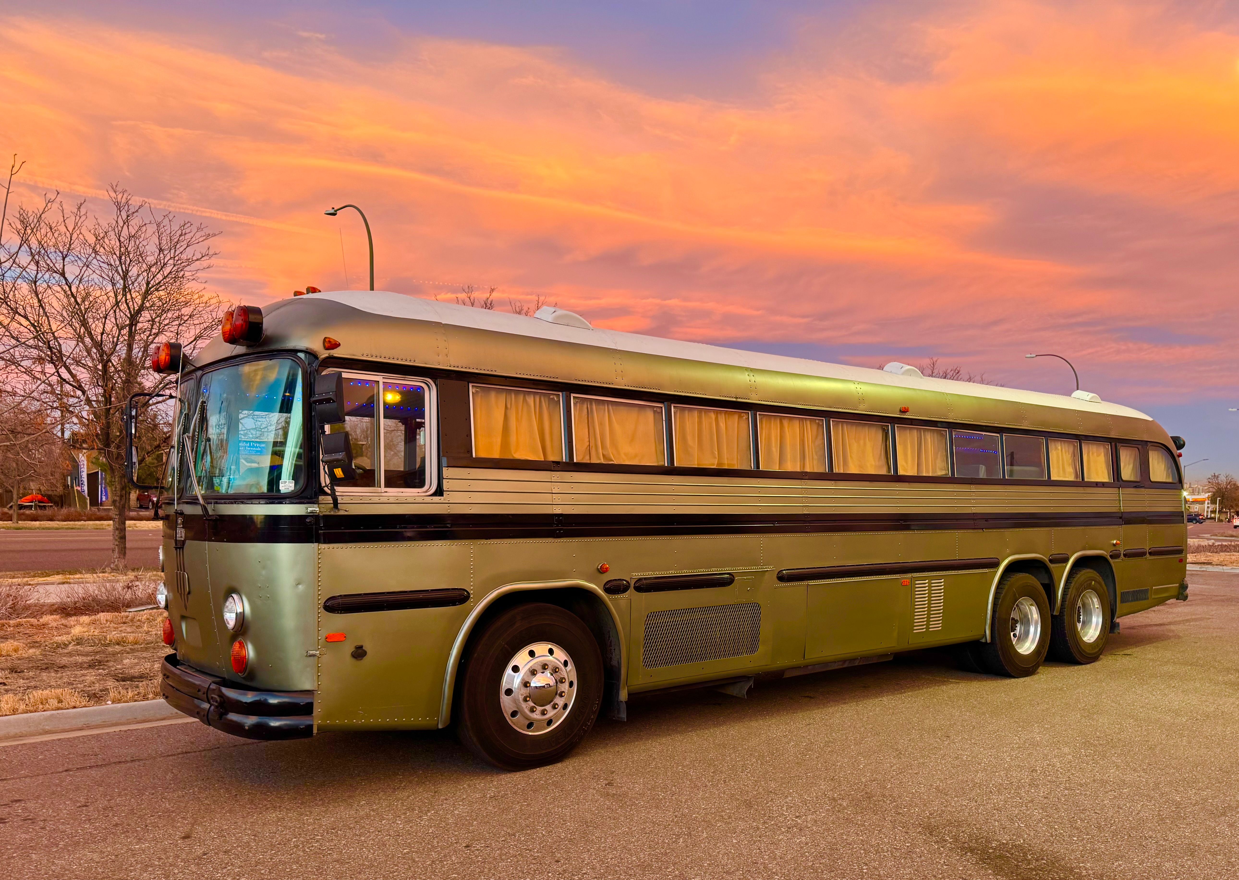 Olive-green vintage coach bus with curtained windows parked on a quiet street at sunset beneath a vivid orange-pink sky, with bare trees and streetlights in the background.