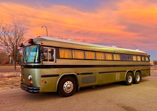 Olive-green vintage coach bus with curtained windows parked on a quiet street at sunset beneath a vivid orange-pink sky, with bare trees and streetlights in the background.
