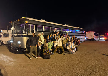 Large group of people posing at night in front of a retro illuminated party bus with purple interior lights parked in a gravel festival parking area.