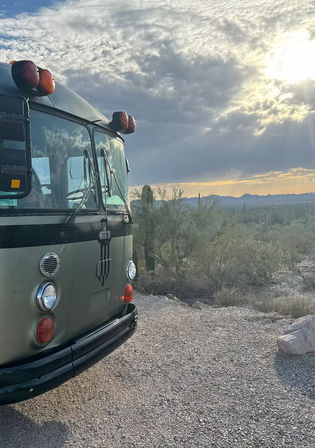Front of a vintage olive-green bus parked on gravel in the southwestern desert with saguaro cacti and sun rays piercing dramatic clouds at sunrise — road‑trip vibe.