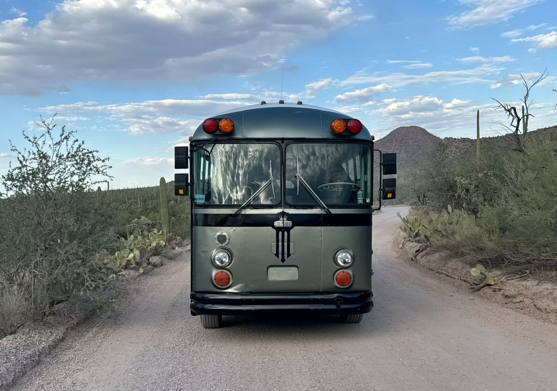Front view of a retro green bus on a dusty desert road lined with saguaro cacti and scrub, low mountains and a partly-cloudy sky creating a road‑trip vibe.