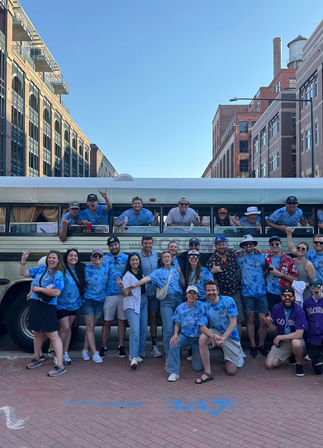 Cheerful large group wearing matching blue tie-dye shirts posing in front of a retro white party bus on a sunny downtown brick-paved street lined with multi-story brick buildings.