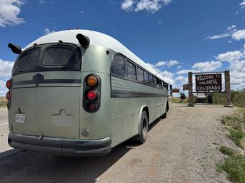 Retro pale-green vintage bus parked beside a roadside 'Welcome to Colorful Colorado' sign under a bright blue sky with scattered clouds