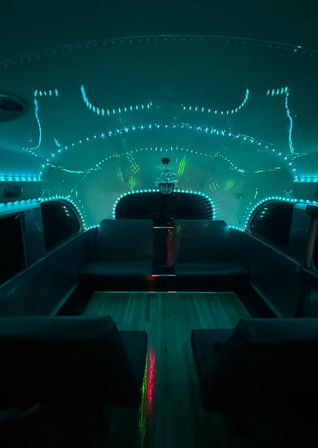 Teal LED-lit retro travel trailer interior with U-shaped bench seating, wood floor and disco-ball light reflections creating a futuristic glamping lounge vibe.