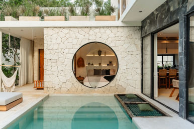 Sunlit modern villa courtyard with turquoise plunge pool, textured stone wall featuring a large circular window revealing a cozy bedroom, hammock lounge and open-plan dining area.