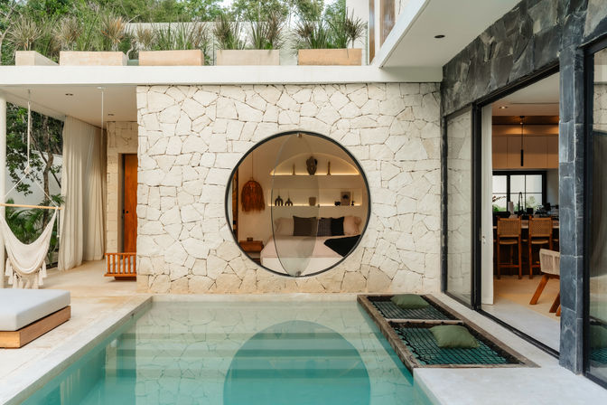 Sunlit modern villa courtyard with turquoise plunge pool, textured stone wall featuring a large circular window revealing a cozy bedroom, hammock lounge and open-plan dining area.