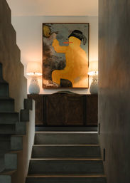 Modern entryway with concrete stairs leading to a wooden sideboard topped by twin pineapple table lamps and a large whimsical yellow painting, warm ambient lighting