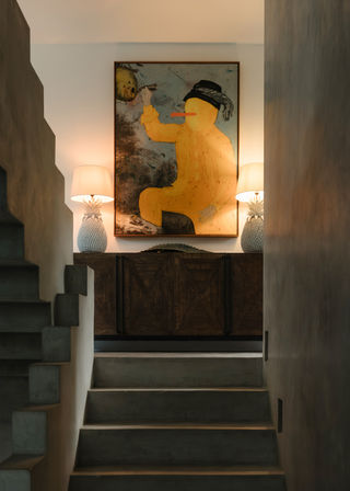 Modern entryway with concrete stairs leading to a wooden sideboard topped by twin pineapple table lamps and a large whimsical yellow painting, warm ambient lighting