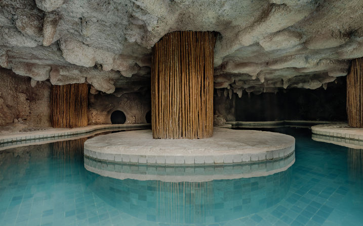 Indoor cave-style pool grotto with turquoise tiled water, circular stone platform surrounding a central wooden-slat column beneath a textured stalactite-like ceiling
