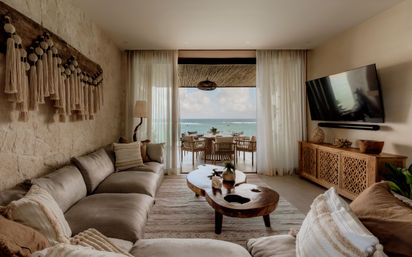 Beachfront living room with neutral boho decor — plush sectional, macramé wall hanging, live-edge wood coffee table, and floor-to-ceiling glass doors opening to a balcony dining set and turquoise ocean view.