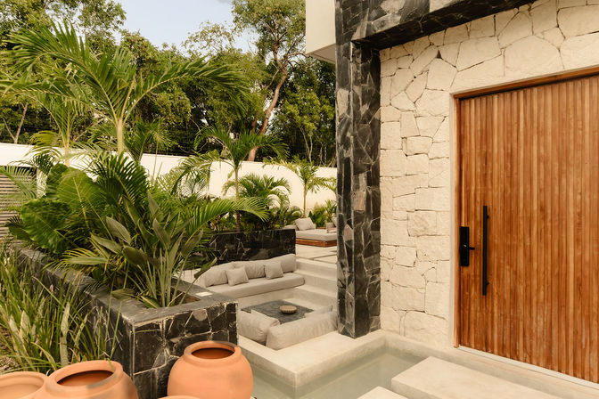 Modern tropical courtyard with a vertical wooden front door, cream stone and dark tile walls, sunken cushioned lounge, shallow water feature, terracotta planters and lush palm greenery