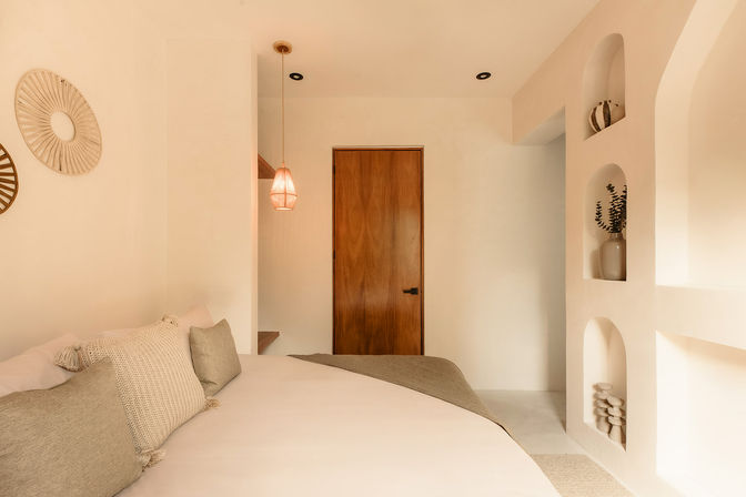 Cozy neutral-toned bedroom with a large bed layered in textured beige and gray pillows, arched built-in wall niches with decorative vases and pottery, a warm pendant light, and a wooden door.