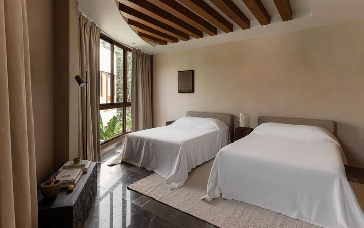 Cozy minimalist twin bedroom with two white-covered beds, large curved floor-to-ceiling window overlooking greenery, wooden ceiling beams and neutral decor