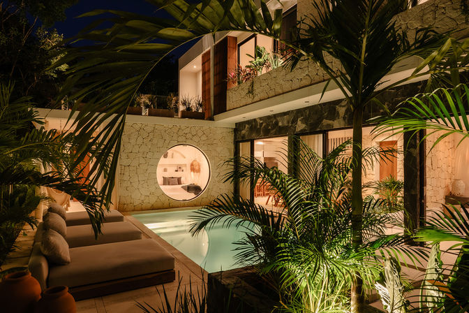 Night view of a tropical modern villa courtyard with an illuminated swimming pool, stone facade and circular window, lush palms and cushioned outdoor loungers.