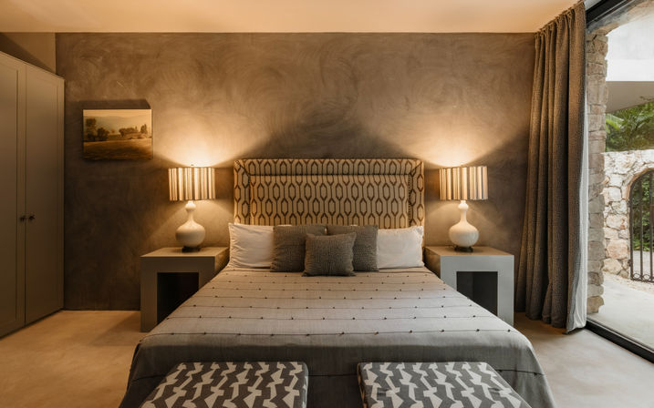 Cozy modern bedroom with patterned headboard, warm-lit bedside lamps, neutral gray walls, patterned benches at the foot and a floor-to-ceiling window revealing a stone archway and garden view.
