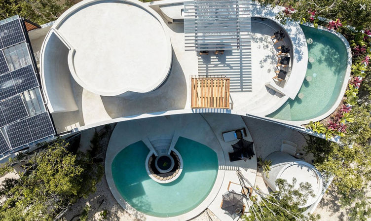 Aerial drone view of a modern curved white concrete coastal villa with solar panels, pergola, two turquoise pools and tropical landscaping with outdoor lounge seating.