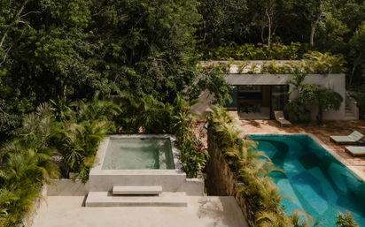 Aerial view of a modern jungle villa surrounded by lush tropical foliage, featuring a concrete plunge pool, a turquoise rectangular swimming pool with sun loungers, stone terrace and rooftop garden.