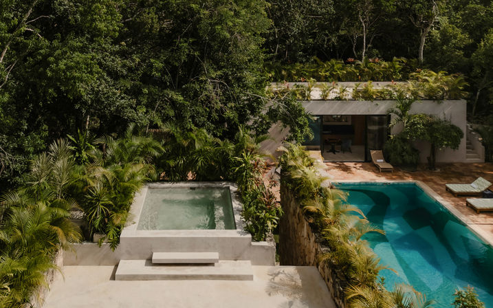 Aerial view of a modern jungle villa surrounded by lush tropical foliage, featuring a concrete plunge pool, a turquoise rectangular swimming pool with sun loungers, stone terrace and rooftop garden.