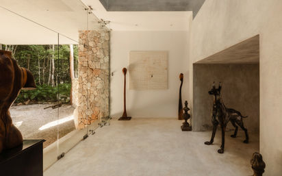 Modern minimalist foyer with floor-to-ceiling glass wall opening to lush forest, exposed stone column, smooth concrete floor, playful bronze dog sculpture, tall wooden totems and grid wall art