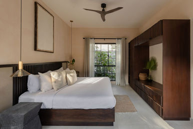 Sunlit modern neutral bedroom with king bed, white linens and textured pillows, dark wood headboard and built-in shelving, large window framing lush greenery, ceiling fan and pendant lights for a minimal tropical-inspired look.
