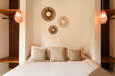Cozy minimalist bedroom with white linens, three textured beige throw pillows, circular rattan wall hangings and warm wicker pendant lights highlighting natural textures.