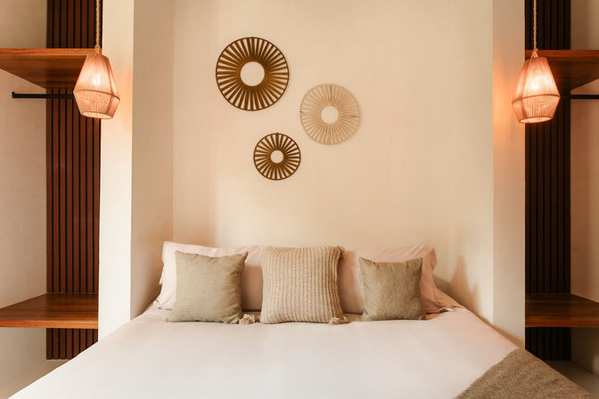 Cozy minimalist bedroom with white linens, three textured beige throw pillows, circular rattan wall hangings and warm wicker pendant lights highlighting natural textures.