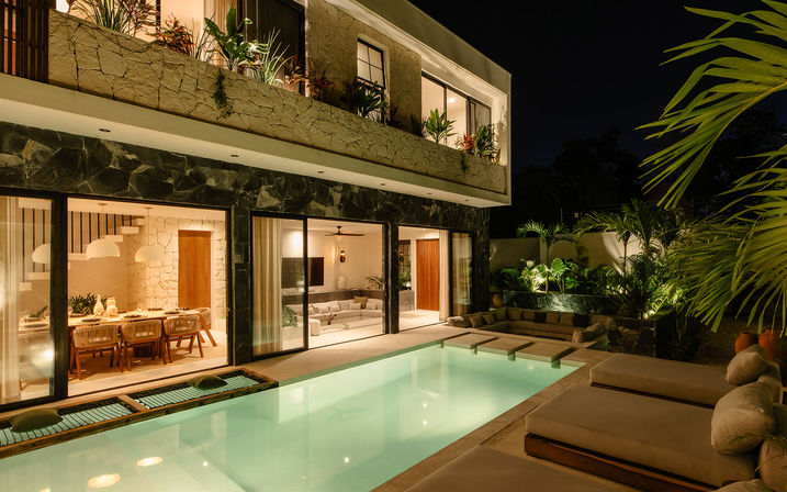 Night view of a modern tropical villa patio with illuminated rectangular pool, cushioned lounge beds, sliding glass doors to cozy living and dining areas, and lush palms